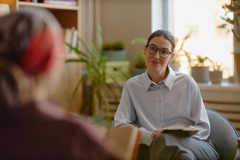 Caucasian young adult woman psychologist wearing glasses listening attentively to unseen patient during therapy session in office, holding clipboard, engaging in professional psychological counseling