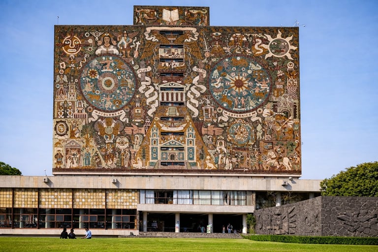 Modern building with large decorative copper relief mural featuring Aztec-inspired designs and symbols on facade above glass windows and green lawn